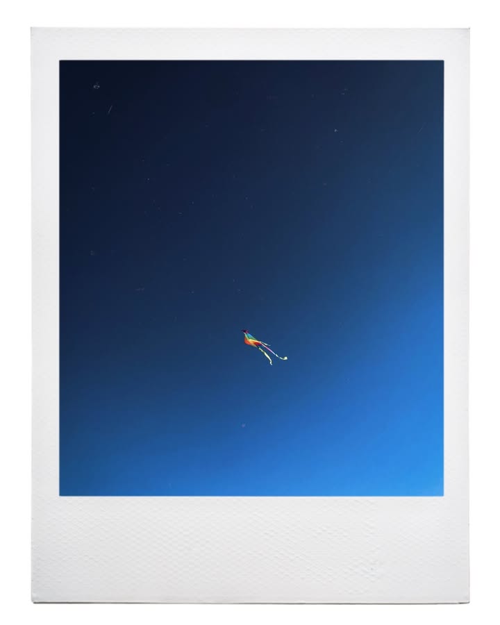 A small multicolored kite with trailing ribbons against a deep blue sky, in an instant-photo-style white frame.